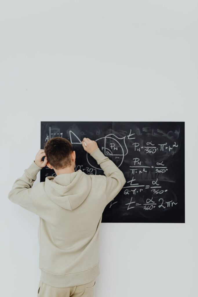 Back view of a teenage boy in a hoodie solving complex math equations on a blackboard indoors.