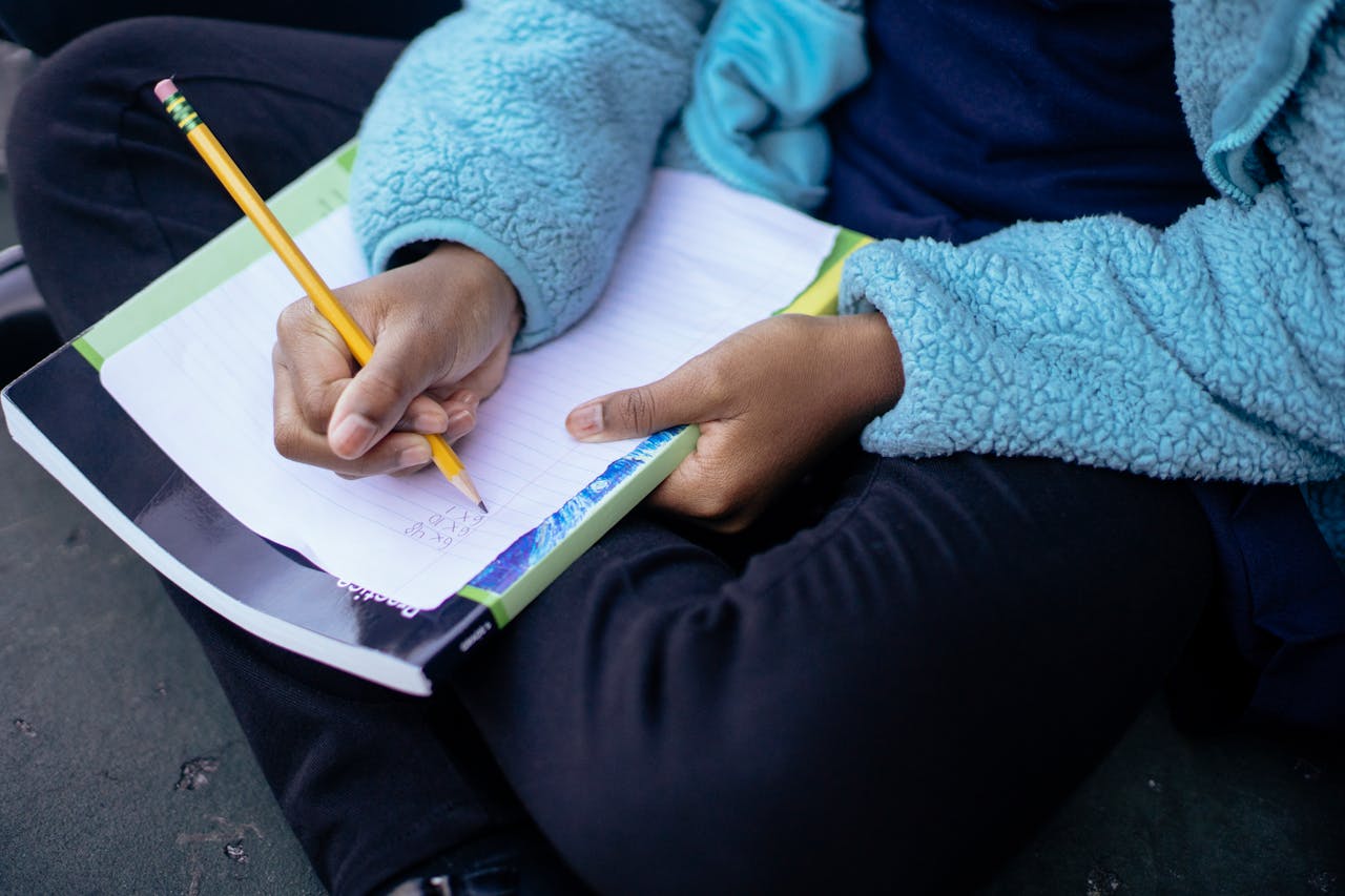 Child sitting outdoors, writing math homework in a notebook with concentration.