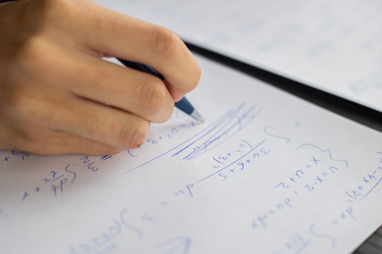 Unrecognizable smart student taking notes on piece of paper while solving mathematical formulas during lesson in classroom on blurred background